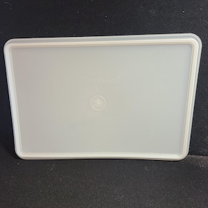 May include: A rectangular, off-white Tupperware lid with a raised rim and a circular indentation in the center. The word "Tupperware" is subtly printed on the lid's surface. The lid is designed to fit a rectangular container.