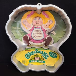 Puede incluir: Un molde para pastel Cabbage Patch Kids vintage con una forma única. El molde presenta un diseño de un Cabbage Patch Kid con cabello rubio y un vestido rosa. Las palabras "Happy Birthday Lauren" están en el vestido. El molde incluye una etiqueta amarilla y verde.