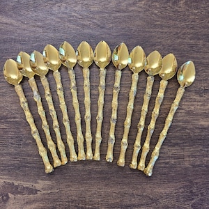 May include: A set of twelve gold-toned spoons arranged in a fan shape. Each spoon features a polished, smooth oval bowl and a textured handle designed to resemble a segmented bamboo stalk, providing a consistent metallic finish throughout.