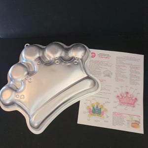 May include: A silver-colored crown-shaped cake pan with a detailed design. The pan is made of metal and has a loop for hanging. A printed instruction sheet for decorating crown cakes is next to the pan.