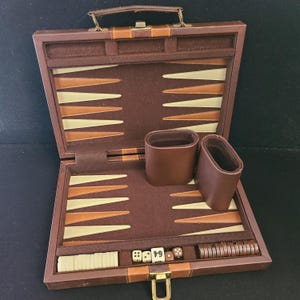May include: Open backgammon set in a brown leather case. The interior features a playing surface with alternating tan and brown triangles. Includes dice, game pieces, and leather dice cups. The case has a handle and a metal clasp.