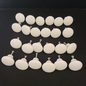 Puede incluir: Una colección de veintiún pomos de armario de cerámica blanca con tornillos plateados. Los pomos son redondos y brillantes, dispuestos sobre una superficie oscura. Adecuados para la restauración de muebles o nuevas construcciones.