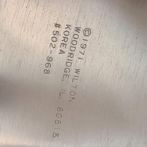 Puede incluir: Primer plano de una superficie met&aacute;lica plateada con texto grabado. El texto incluye "&copy; 1971 WILTON WOODRIDGE, IL. 60515" y "KOREA #502-968". La superficie tiene ara&ntilde;azos visibles.