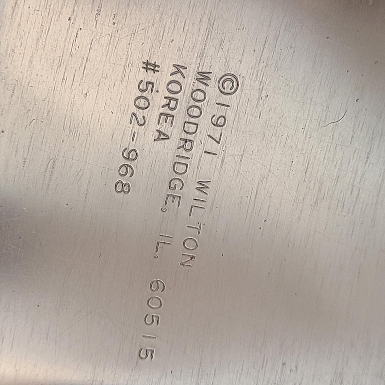 Puede incluir: Primer plano de una superficie met&aacute;lica plateada con texto grabado. El texto incluye "&copy; 1971 WILTON WOODRIDGE, IL. 60515" y "KOREA #502-968". La superficie tiene ara&ntilde;azos visibles.