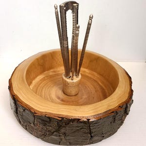 May include: A wooden nut bowl with a natural edge, featuring a central wooden holder containing several metal nutcrackers and picks. The bowl's interior is light brown, contrasting with the darker, textured bark edge.