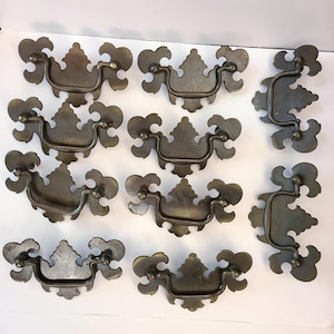 May include: A collection of nine metal drawer pulls featuring a dark, weathered finish. Each pull consists of a decorative backplate with ornate, scrolling edges and a matching swinging bail handle attached by two small, rounded metal posts.