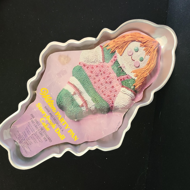 Puede incluir: Un pastel con forma de mu&ntilde;eca en una bandeja plateada. El pastel tiene cabello naranja, un vestido rosa con lunares blancos y mangas y piernas a rayas verdes y blancas. La bandeja incluye una inserci&oacute;n de papel con texto.