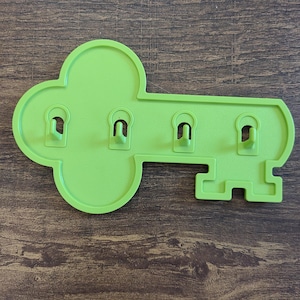 May include: A lime green wall-mounted key holder shaped like a large skeleton key. The flat, plastic-like object features four small, integrated hooks extending outward from the center, designed for hanging items in a horizontal orientation.