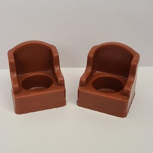 May include: Two brown plastic toy chairs with rounded backs and circular openings in the seats. The chairs are a solid color and appear to be vintage toys. The chairs are side by side on a white surface.