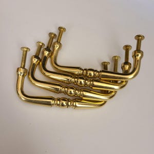 Puede incluir: Un juego de seis tiradores dorados para armarios. Cada tirador tiene un asa curvada con detalles decorativos y un tornillo roscado para el montaje. Los tiradores están dispuestos en un patrón ligeramente superpuesto, mostrando su diseño.