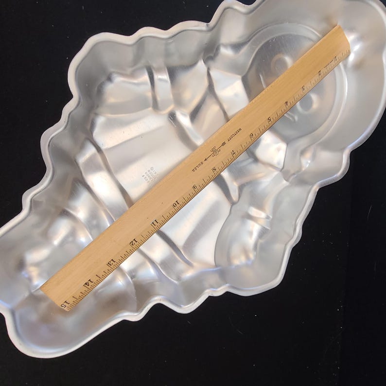 Puede incluir: Un molde para pasteles de aluminio plateado con forma de concha. Una regla de madera, marcada en pulgadas, descansa sobre el molde. El molde tiene un borde festoneado y detalles interiores.