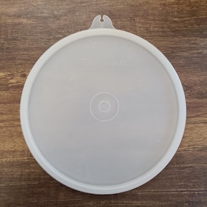 May include: A round, semi-translucent white plastic Tupperware lid featuring a small tab at the top edge for easy removal. The center of the lid displays a circular embossed design, and the brand name Tupperware is clearly visible at the top.