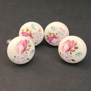 Puede incluir: Cuatro pomos de armario de cerámica blanca con diseños florales. Cada pomo presenta un tulipán rosa, flores de lavanda y detalles dorados y turquesas. Los pomos son redondos y tienen un tornillo plateado.
