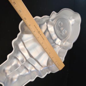 Puede incluir: Un molde para pasteles plateado con forma de cono de helado con un dise&ntilde;o de cara decorativa. Una regla de madera, marcada en cent&iacute;metros, descansa diagonalmente sobre el molde. El molde es de metal y est&aacute; sobre una superficie negra.