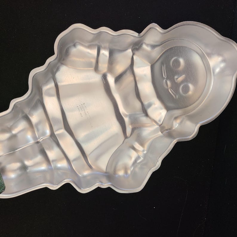 Puede incluir: Un molde para hornear de aluminio plateado con forma de momia. El molde tiene un dise&ntilde;o detallado de cara y cuerpo, con una superficie texturizada. El molde est&aacute; dise&ntilde;ado para hornear pasteles u otras delicias.