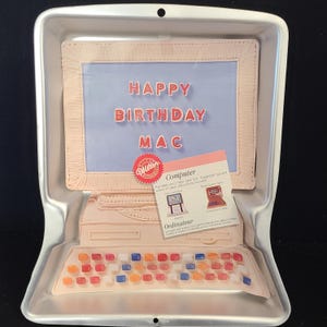 May include: A silver-colored computer-shaped cake pan with a keyboard, screen, and base. The screen displays "HAPPY BIRTHDAY MAC" in red letters. A small card with instructions is included. The pan is designed for baking a cake.