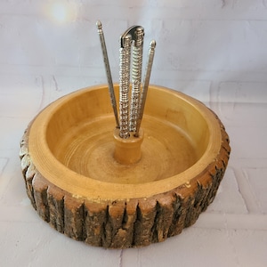 May include: A round wooden bowl with a natural bark exterior and a smooth interior. A central wooden post holds three upright silver-toned metal nut-cracking tools with textured, ridged handles, arranged vertically in the center of the bowl.