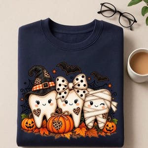 Op de afbeelding: Een marineblauw sweatshirt met een afbeelding van drie cartoon-tanden verkleed voor Halloween. Eén tand draagt een heksenhoed, één heeft een strik met stippen en één is ingewikkeld als een mummie. Ze worden omringd door pompoenen en bladeren.