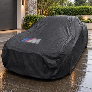 Puede incluir: Una funda de coche negra que cubre un vehículo, con el logotipo M en azul, morado y rojo en el capó. La tela está cubierta de pequeñas gotas de agua, lo que indica un entorno lluvioso, y se ajusta a los contornos de la forma del coche.