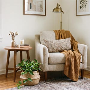 May include: A beige upholstered armchair with wooden legs, draped with a textured mustard yellow knit throw blanket and a rectangular patterned lumbar pillow. Beside it sits a round wooden side table holding a small vase, candle, and books.