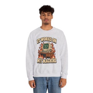 May include: A light grey crewneck sweatshirt featuring a graphic of a smiling computer on a desk surrounded by flames. The text above and below the illustration reads IT WORKS ON MY MACHINE in a bold, retro-style font with dark outlines.