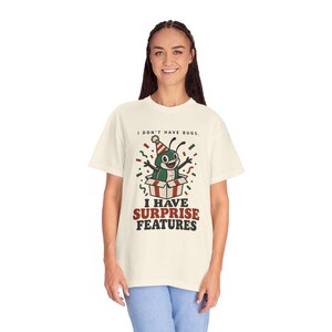 Peut inclure: Un t-shirt à manches courtes couleur crème avec un motif de personnage insecte vert portant un chapeau de fête sortant d'une boîte cadeau. Le texte indique I DON'T HAVE BUGS. I HAVE SURPRISE FEATURES en noir et rouge avec des confettis.