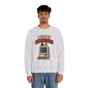 May include: A light grey crewneck sweatshirt featuring a graphic of a vintage computer with a small Christmas tree and a rubber duck wearing a Santa hat on top. The text above the computer reads MERRY COMMITMAS in red and beige.