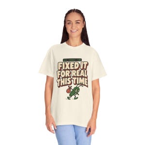 May include: A cream-coloured short-sleeve t-shirt featuring the text "git commit -m" and "FIXED IT FOR REAL THIS TIME" in bold brown letters. Below the text is a green cartoon bug carrying a small red and white polka-dot bundle.