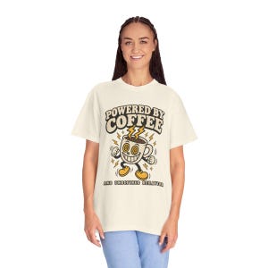 May include: A cream-colored t-shirt featuring a graphic of a cartoon coffee mug with a wide smile, spiral eyes, and yellow lightning bolts. The text reads "POWERED BY COFFEE" at the top and "AND UNDEFINED BEHAVIOR" at the bottom in dark lettering.