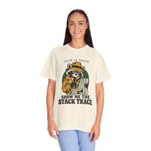 May include: A cream-coloured short-sleeve t-shirt featuring a graphic of a raccoon detective wearing a tan trench coat and fedora, holding a magnifying glass over a long document. The text reads: TALK IS CHEAP. SHOW ME THE STACK TRACE.
