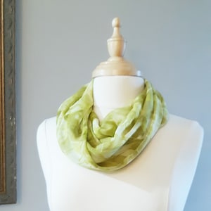 May include: A green and white patterned scarf is displayed on a white mannequin. The scarf is made of a soft, sheer fabric and is tied in a loose knot around the mannequin's neck.