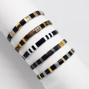 Peut inclure: Cinq bracelets en mosaïque noire, blanche, dorée et argentée. Les bracelets sont empilés les uns sur les autres.