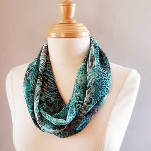 May include: A teal and white patterned infinity scarf with a leopard print design. The scarf is displayed on a wooden mannequin.