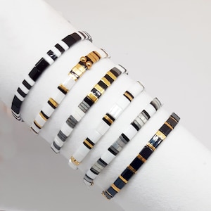 May include: A set of six black, white, and gold beaded bracelets. The bracelets are stacked on top of each other, with the black and white bracelets at the bottom and the gold bracelets at the top.