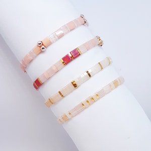 May include: Four pink and white beaded bracelets with gold and silver accents. The bracelets are stacked on top of each other on a white surface.