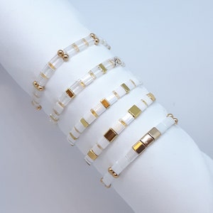 Könnte beinhalten: Fünf weiße und goldene Perlenarmbänder, die auf einer weißen Oberfläche gestapelt sind. Die Armbänder bestehen aus kleinen quadratischen Perlen und goldenen Akzenten.