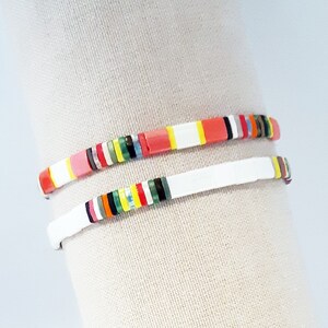 May include: Two colourful bracelets made of small, square beads. The top bracelet has a mix of red, orange, yellow, green, blue, and black beads. The bottom bracelet has white beads with a thin stripe of colour around each bead.