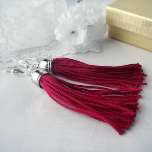 Puede incluir: Un par de pendientes de borla roja con ganchos plateados y una cuenta plateada en la parte superior de cada borla.