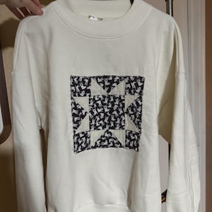 May include: A cream-coloured crewneck sweatshirt featuring a square patch on the chest. The patch displays a black and white star pattern with a repeating ghost motif. The sweatshirt has long sleeves, a ribbed collar, and ribbed cuffs at the hem.