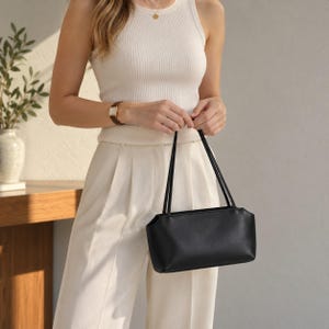 Könnte beinhalten: Eine kleine, rechteckige schwarze Handtasche mit glatter, matter Oberfläche und zwei dünnen, passenden schwarzen Schulterriemen. Die Tasche hat ein strukturiertes Design mit klaren Linien und einem sicheren Verschluss an der Oberseite.