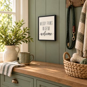 May include: A square wall sign with a black frame and white background featuring the black text MUDDY PAWS WARM welcome. The sign is mounted on a sage green paneled wall next to a green rope leash, a textured towel, and a woven basket.