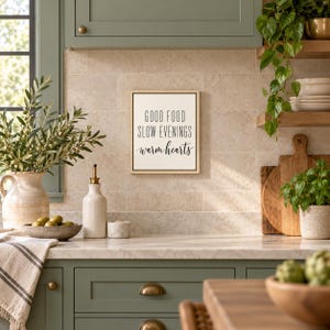 May include: A rectangular wall sign with a light wood frame and a white background featuring the black text: GOOD FOOD SLOW EVENINGS warm hearts. The sign is mounted on a beige tiled wall above a kitchen counter with various items.