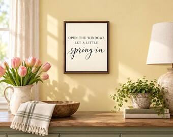 May include: A square wall sign with a dark brown frame and a white background featuring the black text: OPEN THE WINDOWS LET A LITTLE spring in. The sign is displayed above a wooden surface with a vase of pink tulips and a green plant.