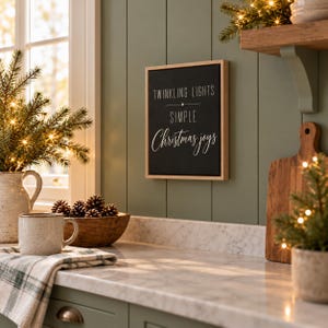 May include: A rectangular wall sign with a light wood frame and a black background featuring the white text TWINKLING LIGHTS, SIMPLE, and Christmas joys, displayed on a green paneled wall above a marble countertop.