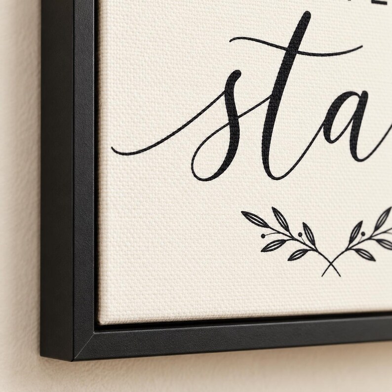 May include: A framed canvas wall art piece featuring a cream-colored background with black cursive text that includes the letters sta and a black line drawing of two symmetrical leafy branches at the bottom, enclosed in a thin black frame.