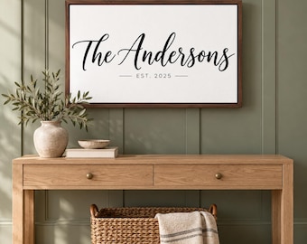 May include: A rectangular wall sign with a dark wood frame featuring the black text The Andersons and EST. 2025 on a white background. The sign is displayed above a light wood console table with two drawers and a woven basket underneath.