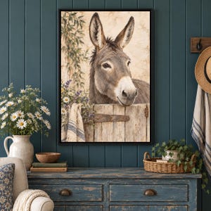 May include: A framed wall art print featuring a brown donkey peering over a weathered wooden fence. The scene includes a textured beige wall, green foliage, and a small cluster of white and purple flowers draped over the fence edge.