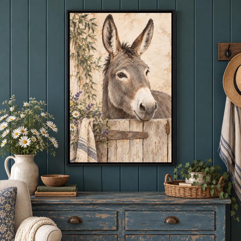 May include: A framed wall art print featuring a brown donkey peering over a weathered wooden fence. The scene includes a textured beige wall, green foliage, and a small cluster of white and purple flowers draped over the fence edge.