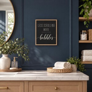 May include: A rectangular wall sign with a light wood frame and a black background. The text in white reads LESS SCROLLING MORE bubbles, with the word bubbles written in a cursive script font. The sign is mounted on a dark blue wall.