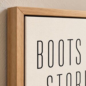 May include: A rectangular canvas wall art piece featuring black text on a white background, framed in a light-toned natural wood. The visible text reads BOOTS and the beginning of the word STORY in a clean, sans-serif black typeface.